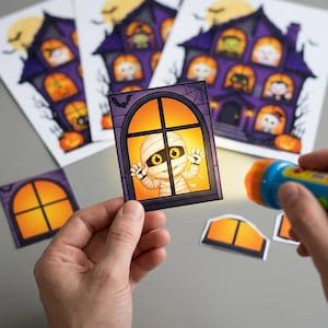 Puede incluir: Una tarjeta rectangular que muestra una momia de dibujos animados tras una ventana con un resplandor naranja, sostenida frente a una linterna. Al fondo, varias hojas impresas muestran una casa encantada morada con personajes y calabazas.