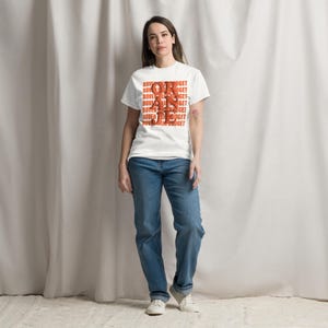 Pode incluir: Uma camiseta branca com as palavras "BOVEN BUDGET" empilhadas verticalmente em letras laranja. A pessoa está usando jeans azuis e tênis brancos. O design da camisa está centralizado na frente.