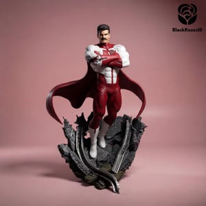 Op de afbeelding: Een beeldje van een superheld in een rood en wit kostuum met een rode cape, staand op een grijze, rotsachtige basis. Het figuur heeft de armen over elkaar. De achtergrond is effen roze. Verzamelaarsobject.