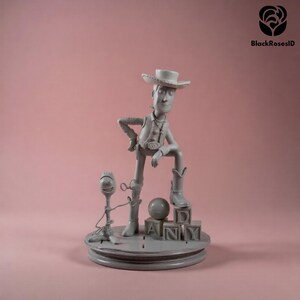 K&ouml;nnte beinhalten: Graue Figur von Woody aus Toy Story, posiert mit einem Bein auf Bl&ouml;cken, die "ANDY" buchstabieren. Eine kleinere Forky-Figur steht in der N&auml;he. Der Sockel ist rund und der Hintergrund ist einfarbig rosa.