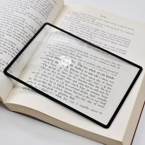 Könnte beinhalten: Eine rechteckige Lupe mit schwarzem Rahmen liegt auf einem offenen Buch. Die klare Linse vergrößert den Text und verbessert die Lesbarkeit. Ideal für das Lesen kleiner Schrift.
