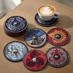 Könnte beinhalten: Eine Sammlung runder Untersetzer mit Wikingerschild-Designs. Die Untersetzer sind in Blau-, Rot- und Hellblautönen gehalten, jeder mit einer anderen Wikinger-Illustration. Eine Kaffeetasse steht auf einem passenden Untersetzer.
