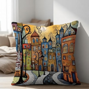 Könnte beinhalten: Quadratisches Zierkissen mit einer skurrilen Illustration einer farbenfrohen Stadtlandschaft. Das Kunstwerk zeigt Gebäude in Orange, Blau und Grün, mit einer gewundenen Kopfsteinpflasterstraße und einem warmen, gelben Himmel.