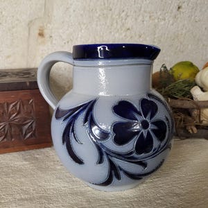 Peut inclure: Une cruche en céramique bleu clair avec un motif floral bleu foncé. La cruche a un corps arrondi, une anse et un bord bleu foncé. Le motif floral comprend une grande fleur et des feuilles tourbillonnantes.