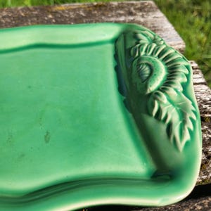 Peut inclure: Un plateau rectangulaire en céramique verte avec un motif floral en relief sur un coin. Le plateau a une finition brillante et un bord légèrement incurvé. Le motif floral présente des feuilles et un élément central circulaire.