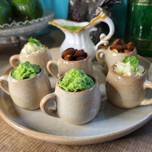 Peut inclure: Un plateau de service en céramique beige contient six petites tasses en céramique tachetées. Les tasses contiennent des trempettes vertes et blanches et des amandes grillées. Une cruche blanche avec une poignée dorée et un bol d'avocats sont en arrière-plan.