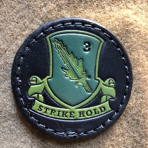 以下が含まれることがあります： 剣と葉が描かれた緑色の盾を持つ黒い革のパッチ。盾には「3」の数字が書かれている。「STRIKE HOLD」という文字が盾の下の緑色のバナーに書かれている。
