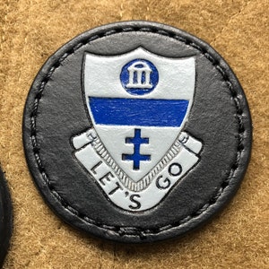 Op de afbeelding: Een zwarte leren patch met een wit schild met een blauw en wit wapenschild met de tekst "Let's Go". Het wapenschild bevat een gebouw met drie pilaren en een kruis.