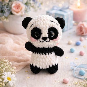 May include: A handmade crochet panda bear with black and white yarn. The panda has black eyes, ears, and limbs, with a white body and face. The panda has pink cheeks and is standing on a white surface.