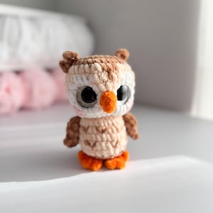 May include: A handmade crochet owl in shades of brown, beige, and orange. The owl has large, expressive eyes, a small orange beak, and tiny feet. The soft, textured yarn creates a cuddly appearance, perfect for a nursery or as a decorative item.