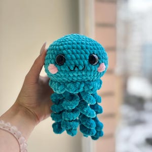 Pode incluir: Um brinquedo de água-viva de crochê turquesa com olhos pretos e bochechas rosadas. O brinquedo de pelúcia tem uma cabeça redonda e tentáculos pendurados. O brinquedo é feito de fio macio e texturizado, projetado para ser um item fofo e aconchegante.
