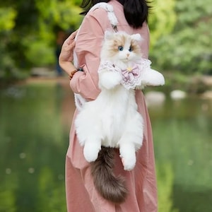 Peut inclure: Un sac à dos en peluche en forme de chat avec une longue fourrure blanche, une queue à pointe brune et des yeux bleus. Le chat porte un col en dentelle blanche avec un petit nœud mauve et un accent central doré. Le sac est muni d'une sangle.