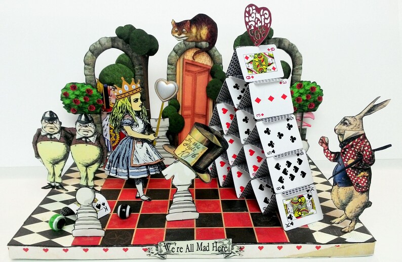 May include: A 3D papercraft scene depicting characters from Alice in Wonderland. Alice stands on a checkered board with a Queen, two Cheshire Cats, and a Mad Hatter. A house of cards stands in the background with the White Rabbit holding a pocket watch.