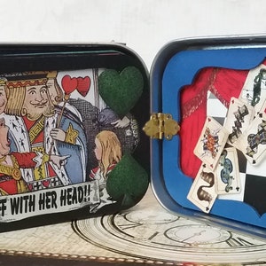 May include: A vintage tin box with a black and white illustration of playing cards and the text "Off with her head!" on the front. The back of the box has a blue frame with a red curtain and a chessboard with playing cards falling from the top.