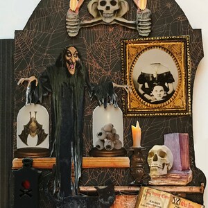 K&ouml;nnte beinhalten: Eine schwarze Katze sitzt vor einer gruseligen Halloween-Szene. Eine Hexe in einem langen schwarzen Gewand h&auml;lt zwei Glaskuppeln, eine mit einer Fledermaus und die andere mit Aug&auml;pfeln. Ein Sch&auml;del sitzt auf einem Regal mit einem Buch mit dem Titel "Sleeping Curses" und "Witch's Brew".