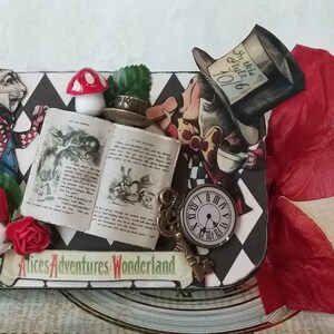 May include: A black and white checkered box with a red ribbon. The box is decorated with images from Alice in Wonderland, including the Mad Hatter, the White Rabbit, and a pocket watch. The text "Alice's Adventures Wonderland" is printed on the box.