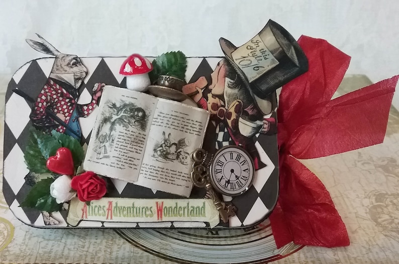 May include: A black and white checkered box with a red ribbon. The box is decorated with images from Alice in Wonderland, including the Mad Hatter, the White Rabbit, and a pocket watch. The text "Alice's Adventures Wonderland" is printed on the box.