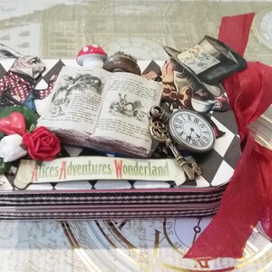 May include: A black and white checkered box with a red ribbon. The box is decorated with images from Alice in Wonderland, including the White Rabbit, the Mad Hatter, a book, a key, and a pocket watch. The box has the text "Alice's Adventures Wonderland" on the front.