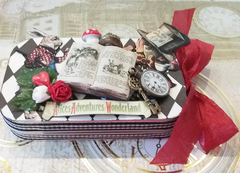 May include: A black and white checkered box with a red ribbon. The box is decorated with images from Alice in Wonderland, including the White Rabbit, the Mad Hatter, a book, a key, and a pocket watch. The box has the text "Alice's Adventures Wonderland" on the front.