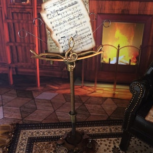 May include: A gold metal music stand with a sheet of music on it. The stand has a decorative design with gears and other mechanical elements. The music stand is in front of a fireplace with a fire burning in it.