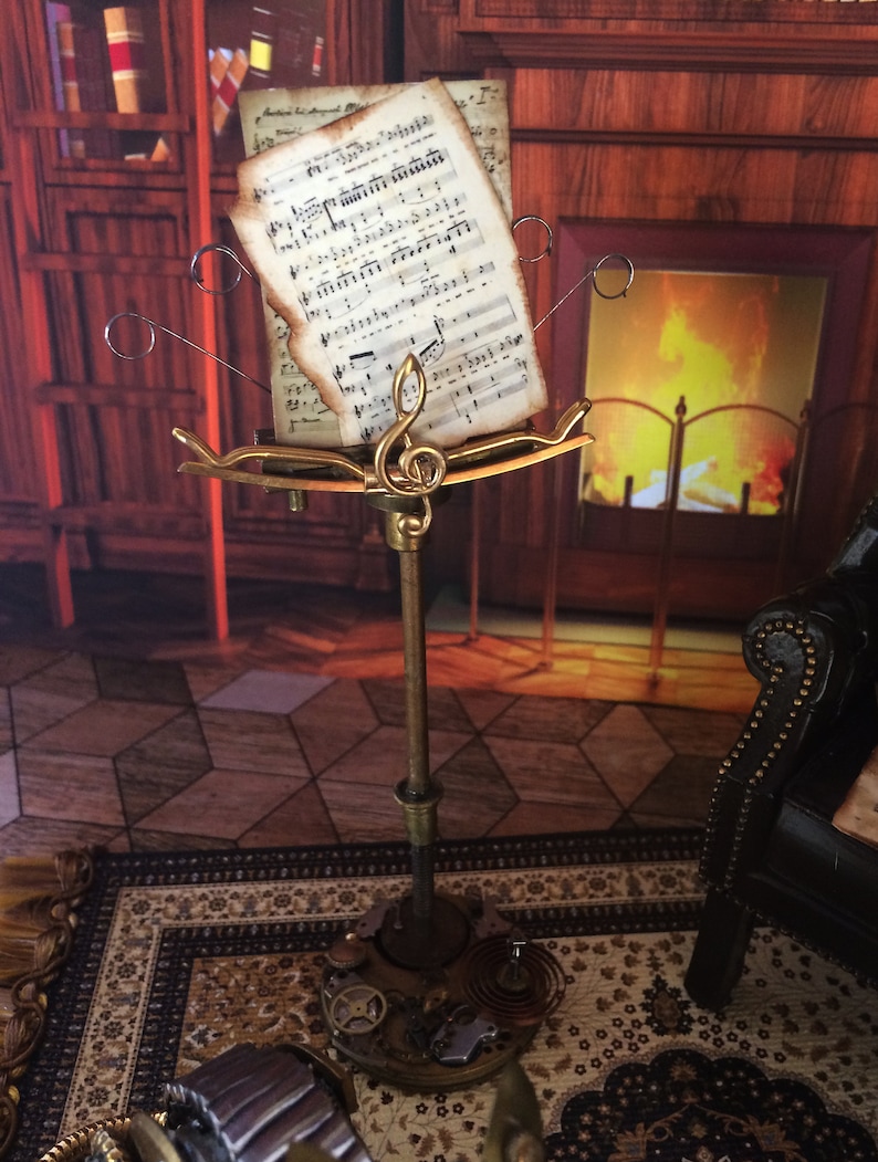 May include: A gold metal music stand with a sheet of music on it. The stand has a decorative design with gears and other mechanical elements. The music stand is in front of a fireplace with a fire burning in it.