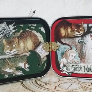 May include: A vintage tin box with a hinged lid, decorated with Alice in Wonderland characters. The front panel features the Cheshire Cat and Alice, with the text "We're All Mad Here". The back panel features the White Rabbit and the Mad Hatter, with the text "Twinkle, Twinkle, Little Bat".