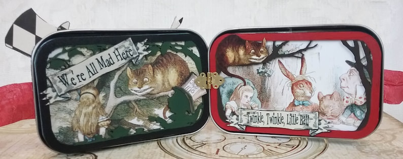 May include: A vintage tin box with a hinged lid, decorated with Alice in Wonderland characters. The front panel features the Cheshire Cat and Alice, with the text "We're All Mad Here". The back panel features the White Rabbit and the Mad Hatter, with the text "Twinkle, Twinkle, Little Bat".