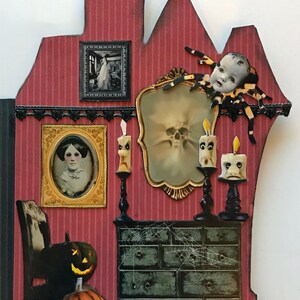K&ouml;nnte beinhalten: Ein rotes und schwarzes Papierhaus mit einem gruseligen Innenraum. Das Haus hat einen schwarzen Stuhl, eine Kommode mit Spinnweben, einen Spiegel, der einen Sch&auml;del reflektiert, und einen gruseligen Puppenkopf mit einer Spinne. Es gibt auch zwei Jack-o'-Lanterns und drei Kerzen mit Gesichtern.