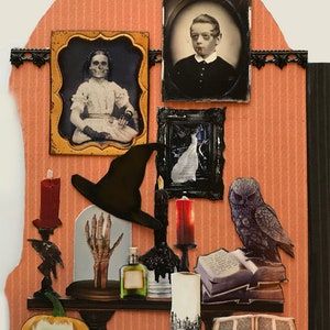 K&ouml;nnte beinhalten: Eine Halloween-Szene mit einem schwarzen Hexenhut, einer skelettartigen Hand in einer Glaskuppel, einem geschnitzten K&uuml;rbis mit einem Sch&auml;delgesicht und Kerzenst&auml;ndern mit roten und schwarzen Kerzen. Au&szlig;erdem gibt es zwei antike gerahmte Portr&auml;ts, ein Buch und eine Eulenfigur.