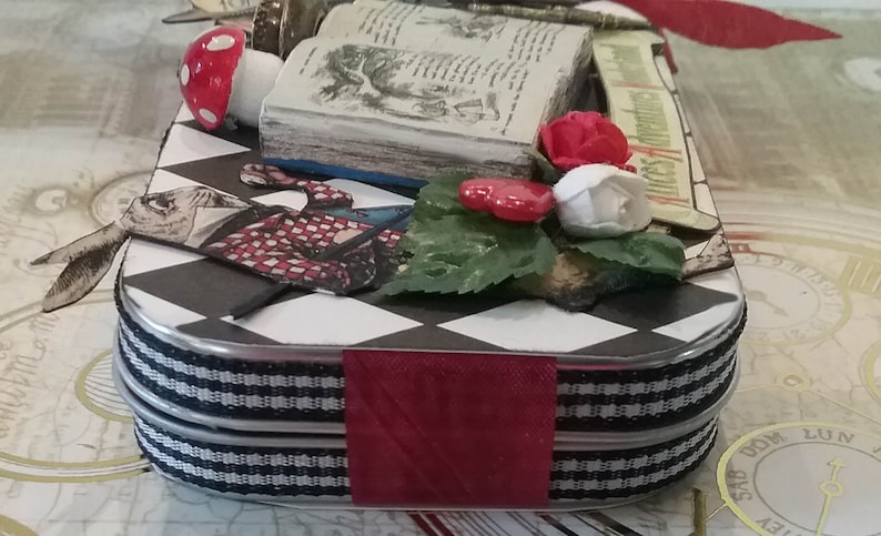 May include: A black and white checkered tin box with a red ribbon and a book, a mushroom, and flowers on top. The book has the title "Alice's Adventures in Wonderland" on the spine.