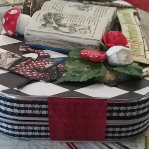 May include: A black and white checkered tin box with a red ribbon and a book, a mushroom, and flowers on top. The book has the title "Alice's Adventures in Wonderland" on the spine.