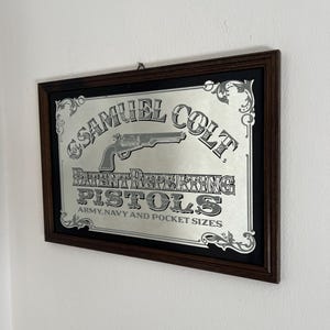 Op de afbeelding: Een rechthoekige wandspiegel in een donkerbruine houten lijst met een zilverkleurige afbeelding van een revolver en de tekst: SAMUEL COLT PATENT REPEATING PISTOLS ARMY, NAVY AND POCKET SIZES. Het ontwerp bevat decoratieve krulmotieven.
