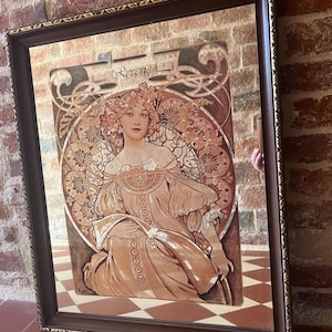 Peut inclure: Une impression d'art encadrée représentant une femme avec des fleurs dans les cheveux et une robe fluide. L'œuvre d'art a un style vintage avec des tons chauds de marron, beige et or. Le mot "Reverie" est visible en haut. Le cadre est marron foncé avec des accents dorés.