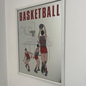 Op de afbeelding: Een rechthoekige wandspiegel met een witte lijst en een grafische illustratie van drie basketballers in rood-grijze uniformen. Het woord BASKETBALL staat in grote rode hoofdletters bovenaan gedrukt, met een basketring en bord zichtbaar.