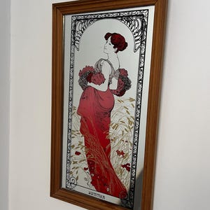 Peut inclure: Un miroir mural rectangulaire dans un cadre en bois présentant une impression décorative d'une personne aux cheveux roux portant une longue robe rouge. La silhouette se tient dans un champ de blé, tenant des fleurs, avec le mot SUMMER en bas.
