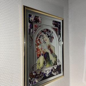 May include: A framed Art Nouveau style print featuring a woman holding flowers. The artwork has a silver background with a gold frame. The woman is wearing a yellow dress and a floral headdress. The print has red, purple, and green accents.