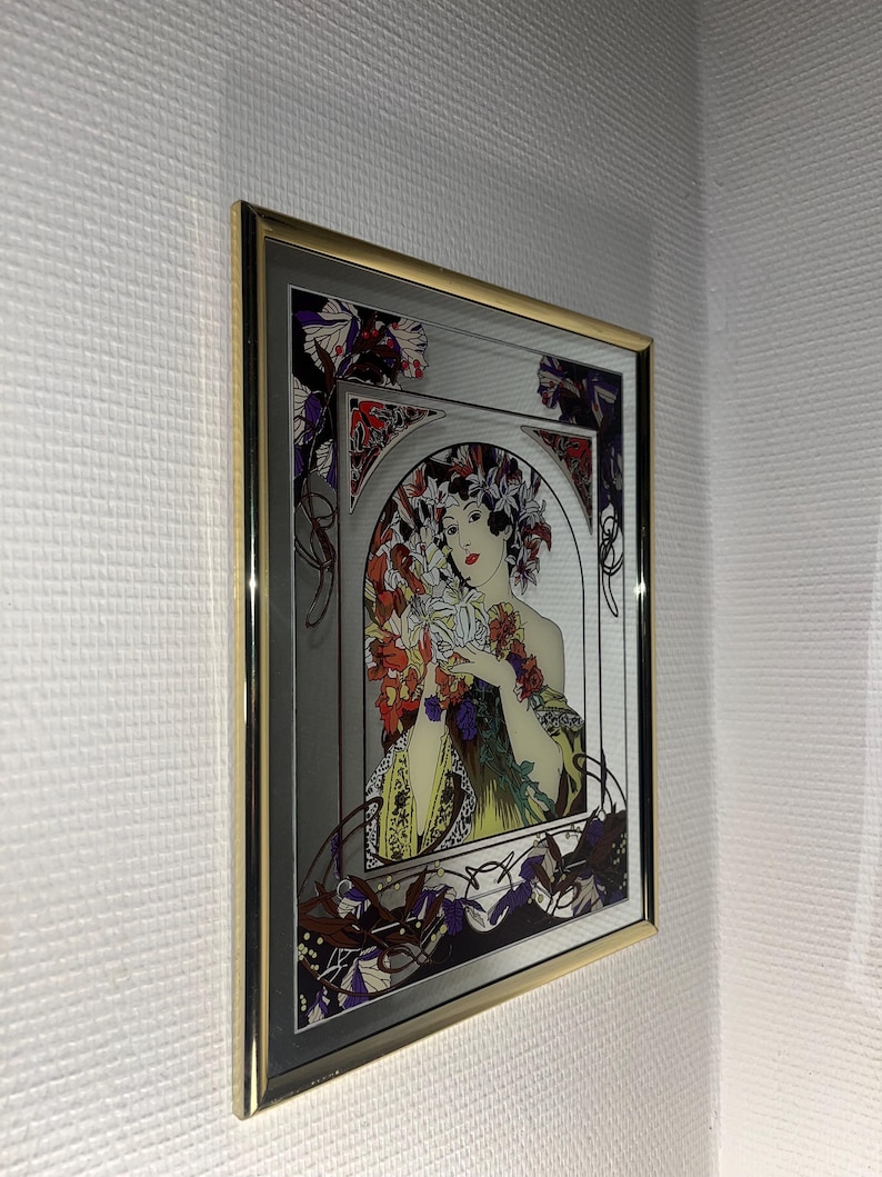 May include: A framed Art Nouveau style print featuring a woman holding flowers. The artwork has a silver background with a gold frame. The woman is wearing a yellow dress and a floral headdress. The print has red, purple, and green accents.
