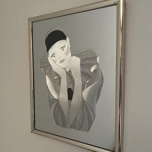 Peut inclure: Un miroir mural carré présentant une illustration imprimée d'une personne avec une casquette noire et un col à volants. Le personnage porte un maquillage stylisé avec des accents en forme de larme sous les yeux. Le miroir est encadré d'un cadre en métal argenté.