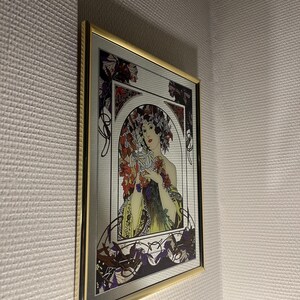 May include: A framed mirror featuring an Art Nouveau style illustration of a woman surrounded by flowers. The artwork has a gold frame and is mounted on a textured white wall. The woman is holding flowers, with a color palette of red, purple, and green.
