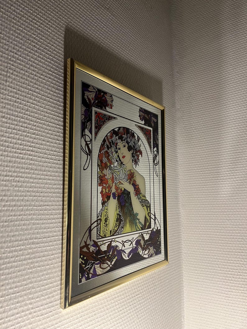May include: A framed mirror featuring an Art Nouveau style illustration of a woman surrounded by flowers. The artwork has a gold frame and is mounted on a textured white wall. The woman is holding flowers, with a color palette of red, purple, and green.