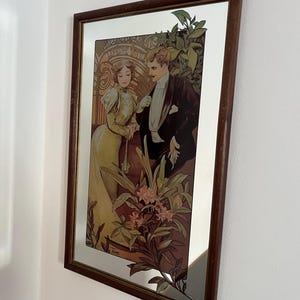Peut inclure: Miroir encadré avec une illustration vintage d'un homme et d'une femme en tenue de soirée. L'œuvre utilise des bruns, des verts et des jaunes, avec des détails floraux. Le cadre est en bois foncé.