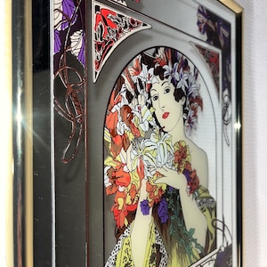 May include: Art Nouveau style framed print featuring a woman with flowers. The artwork has a gold frame and a color palette of red, yellow, purple, and green. The woman is holding a bouquet of flowers.
