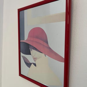 Peut inclure: Impression encadrée d'une femme portant un grand chapeau rouge, avec un cadre rouge. L'œuvre d'art a une esthétique vintage, avec des couleurs douces et une attention particulière au visage et aux épaules de la femme. Le cadre est rouge brillant.