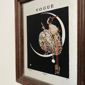 Peut inclure: Une impression encadrée représentant une illustration de couverture du magazine Vogue. L'œuvre montre une silhouette vêtue d'une robe à motifs assise sur un croissant de lune blanc sur fond noir. Le mot VOGUE est imprimé en noir en haut.