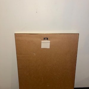 May include: Back of a framed artwork, featuring a brown cardboard backing. A white paper label is affixed near the top, and a metal hanger is visible. The frame's edge is a light brown color. The background is a plain white wall.
