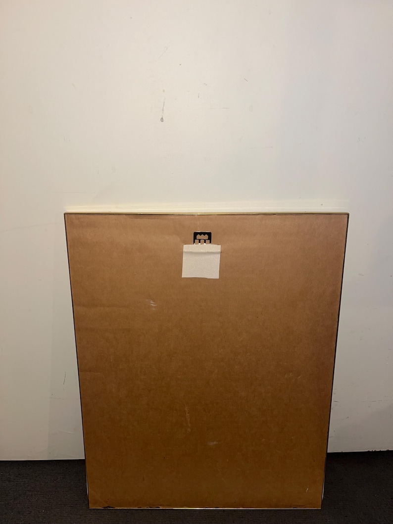 May include: Back of a framed artwork, featuring a brown cardboard backing. A white paper label is affixed near the top, and a metal hanger is visible. The frame's edge is a light brown color. The background is a plain white wall.