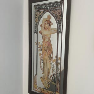 Peut inclure: Un miroir mural rectangulaire orné d'une impression Art Nouveau représentant une personne entourée de fleurs. L'image montre une silhouette en robe dorée fluide avec des détails floraux, encadrée de bois brun foncé. Le texte "Reverie du Soir" est visible.