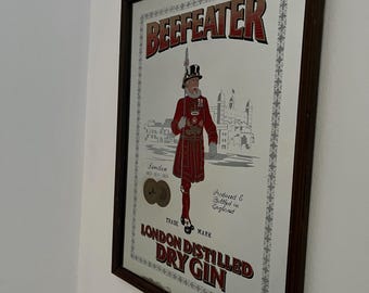 May include: A framed mirror featuring a vintage Beefeater London Dry Gin advertisement. The artwork depicts a Beefeater guard in a red uniform, with the brand name in bold letters. The frame is dark brown.