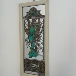 Peut inclure: Un miroir mural rectangulaire avec un cadre couleur crème, orné d'une impression décorative représentant une silhouette en robe verte parmi des arbres. L'œuvre inclut le texte "HIVER" en bas dans une police stylisée et un motif floral circulaire.