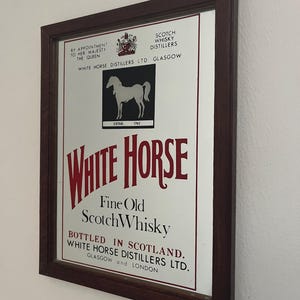 Peut inclure: Un miroir mural rectangulaire dans un cadre en bois brun foncé présentant une publicité pour le whisky White Horse Fine Old Scotch. L'impression comprend un carré noir avec une silhouette de cheval blanc, le texte "ESTAB. 1742" et "BY APPOINTMENT TO HER MAJESTY THE QUEEN."